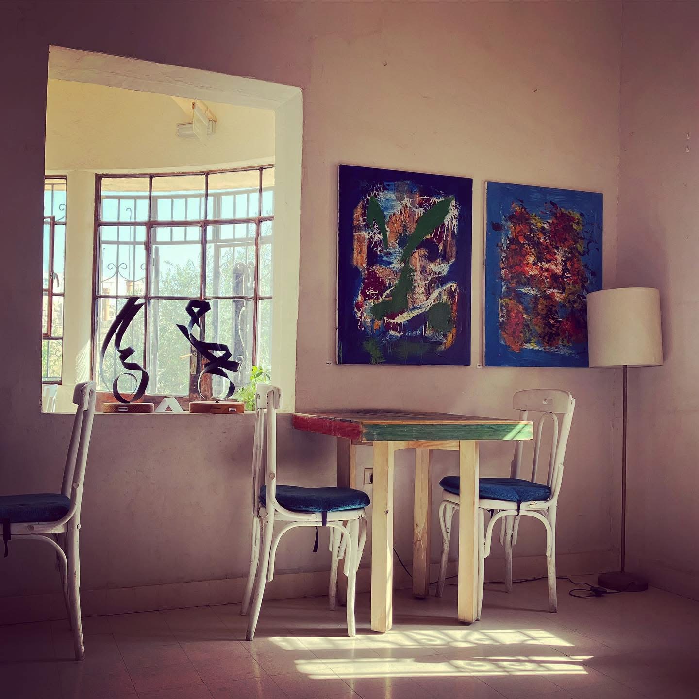Two abstract paintings beside a window with Arabic calligraphy sculptures, above a colorful reclaimed-wood table and white bentwood chairs