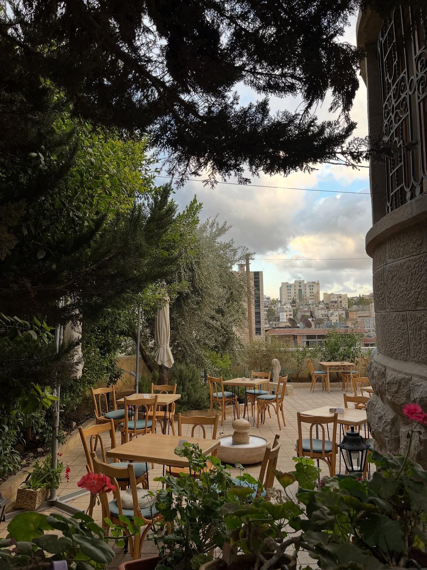 The Fann wa Chai garden at dusk, tables empty under darkening trees with the lights of Amman in the distance