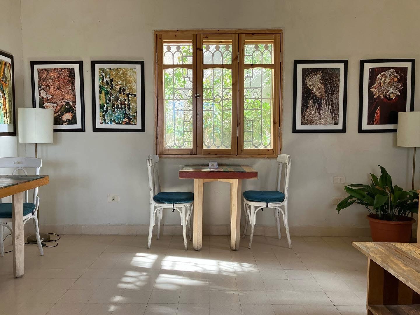 A wall of framed photographic prints hung around a wooden window at Fann wa Chai, with a small table and two white chairs beneath