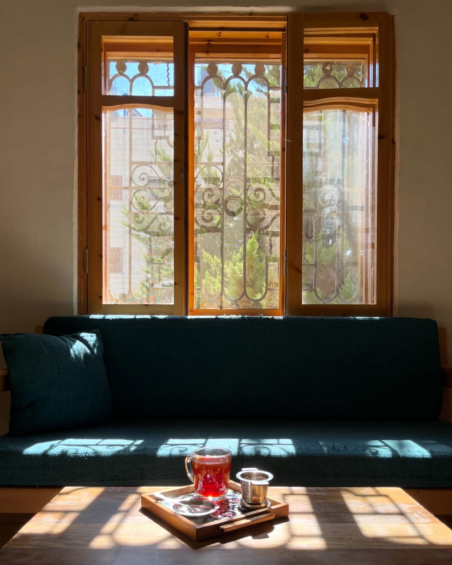 A tray of black tea on a wooden table in front of a deep-teal window seat, with patterned ironwork shadows falling across the cushions